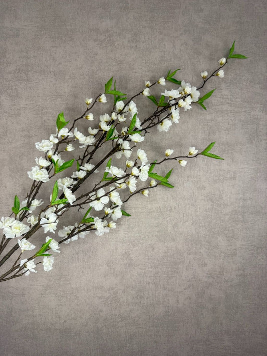 Lange kunsttak met witte kersenbloesem en groene blaadjes tegen een beige achtergrond. Perfect voor een frisse en natuurlijke sfeer in huis.