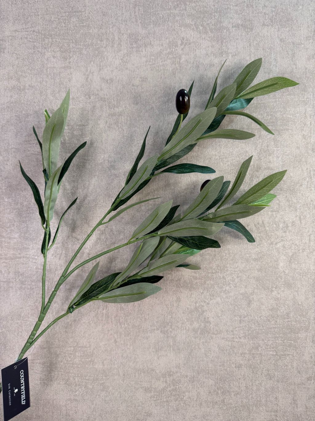 Groene kunsttak met olijfvruchten en langwerpige bladeren tegen een neutrale achtergrond.