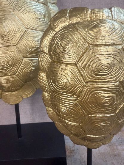 Twee goudkleurige schildvormige ornamenten op zwarte voet, staand op houten ondergrond tegen een neutrale grijze achtergrond.