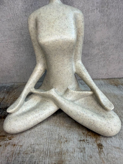 Ornament van een zittend figuur in lotuspositie in beige-grijze kleur op een houten ondergrond.