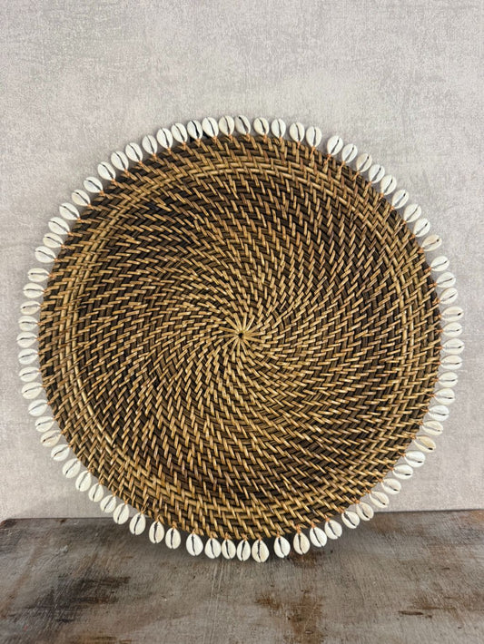 Ronde rotan placemat met witte schelprand, gebruikt als decoratief onderbord of dienblad op houten ondergrond.