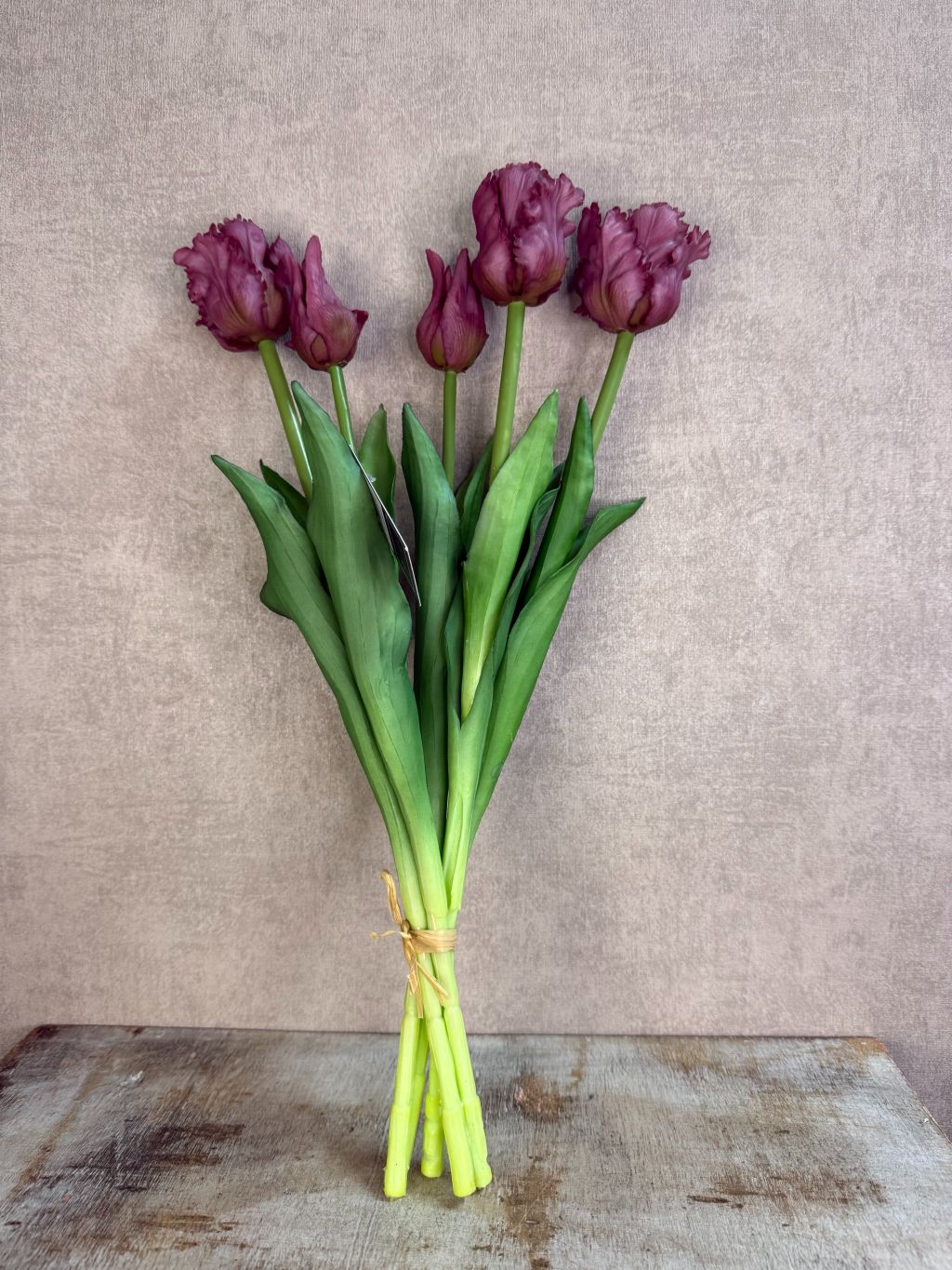 Boeket real touch papegaaien tulpen in paarse kleur met groene bladeren, samengebonden en staand op een houten ondergrond tegen een neutrale achtergrond.
