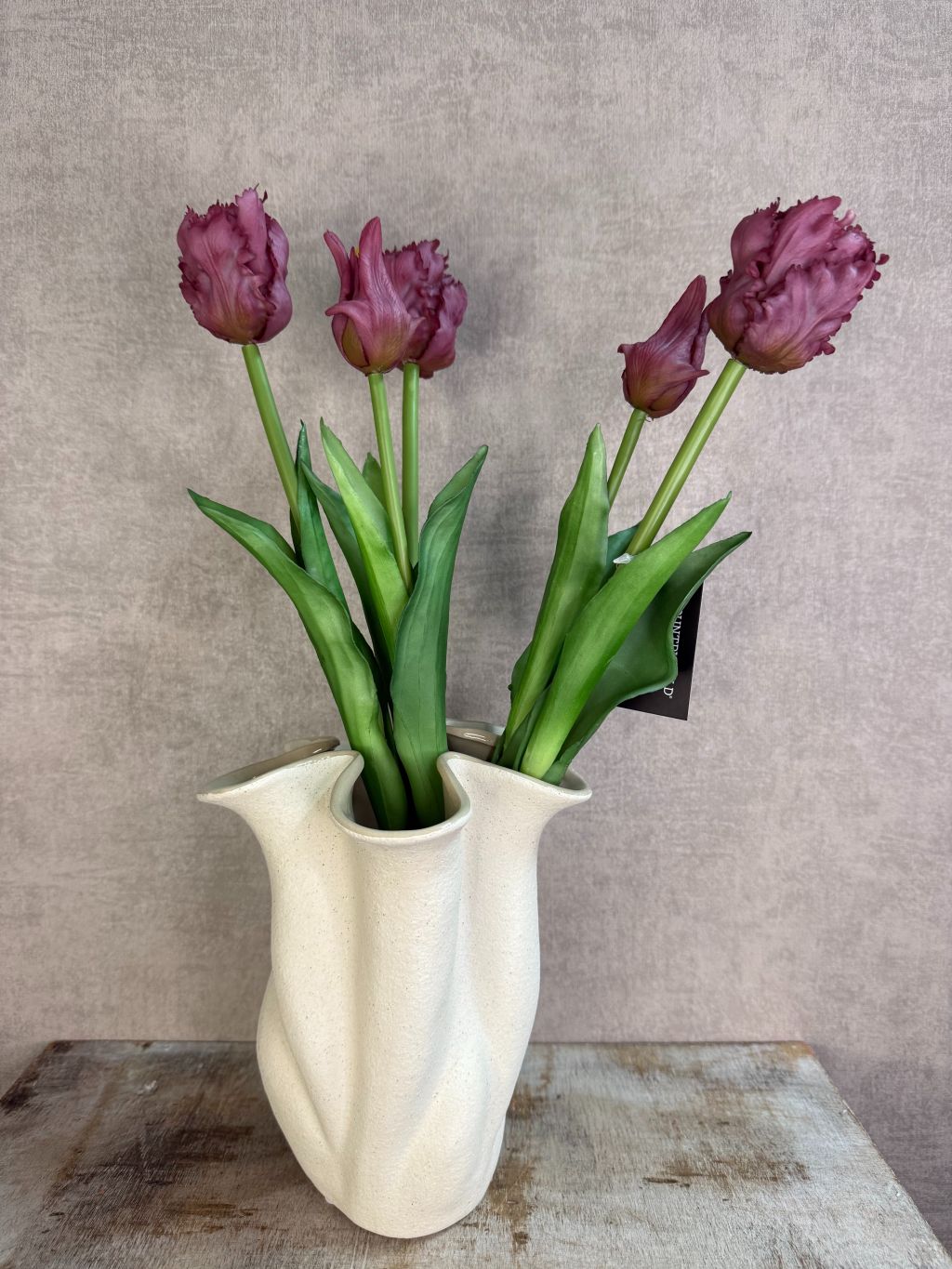 Boeket real touch papegaaien tulpen in paarse kleur met groene bladeren, samengebonden en staand op een houten ondergrond tegen een neutrale achtergrond. met vaas