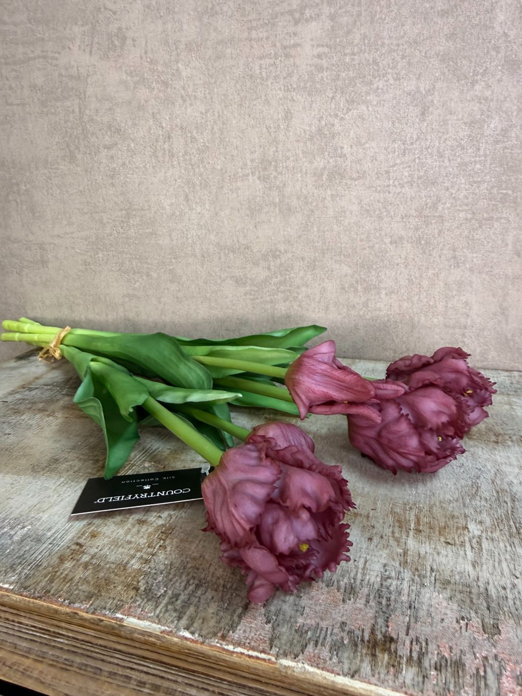 Boeket real touch papegaaien tulpen in paarse kleur met groene bladeren, samengebonden en staand op een houten ondergrond tegen een neutrale achtergrond.