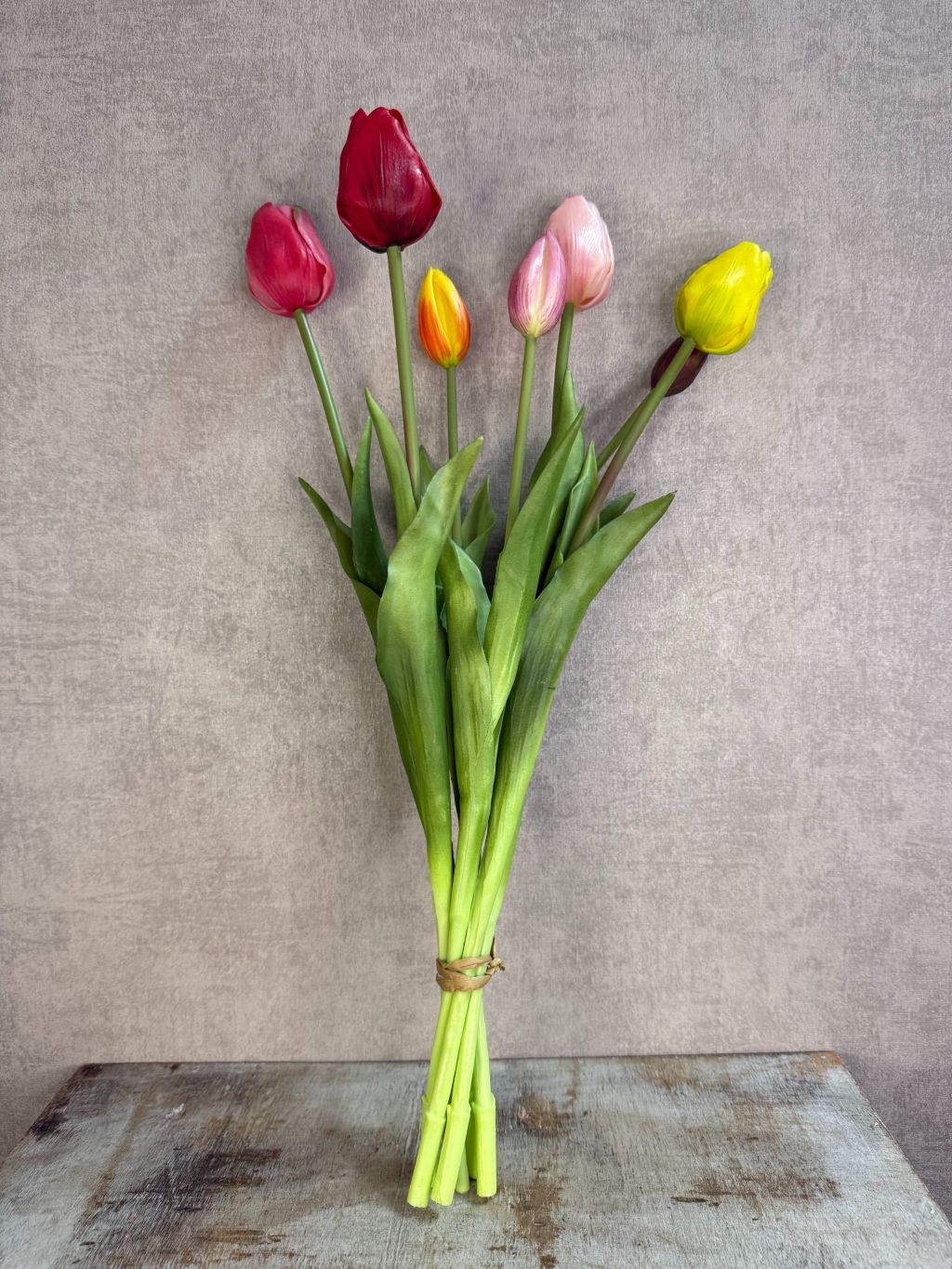 Bos real touch tulpen in een bonte mix van kleuren met groene bladeren, staand op een houten ondergrond tegen een neutrale achtergrond.