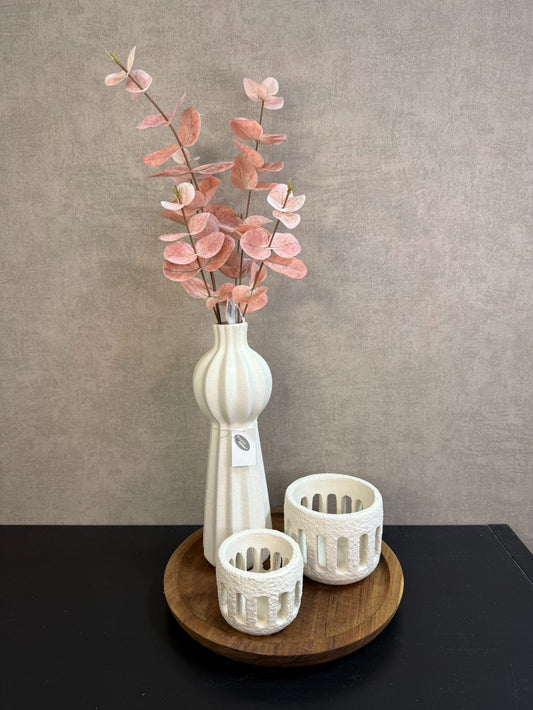 Ronde houten tray met crème vaas met roze kunsttakken en twee crème waxinelichthouders op een zwarte tafel.