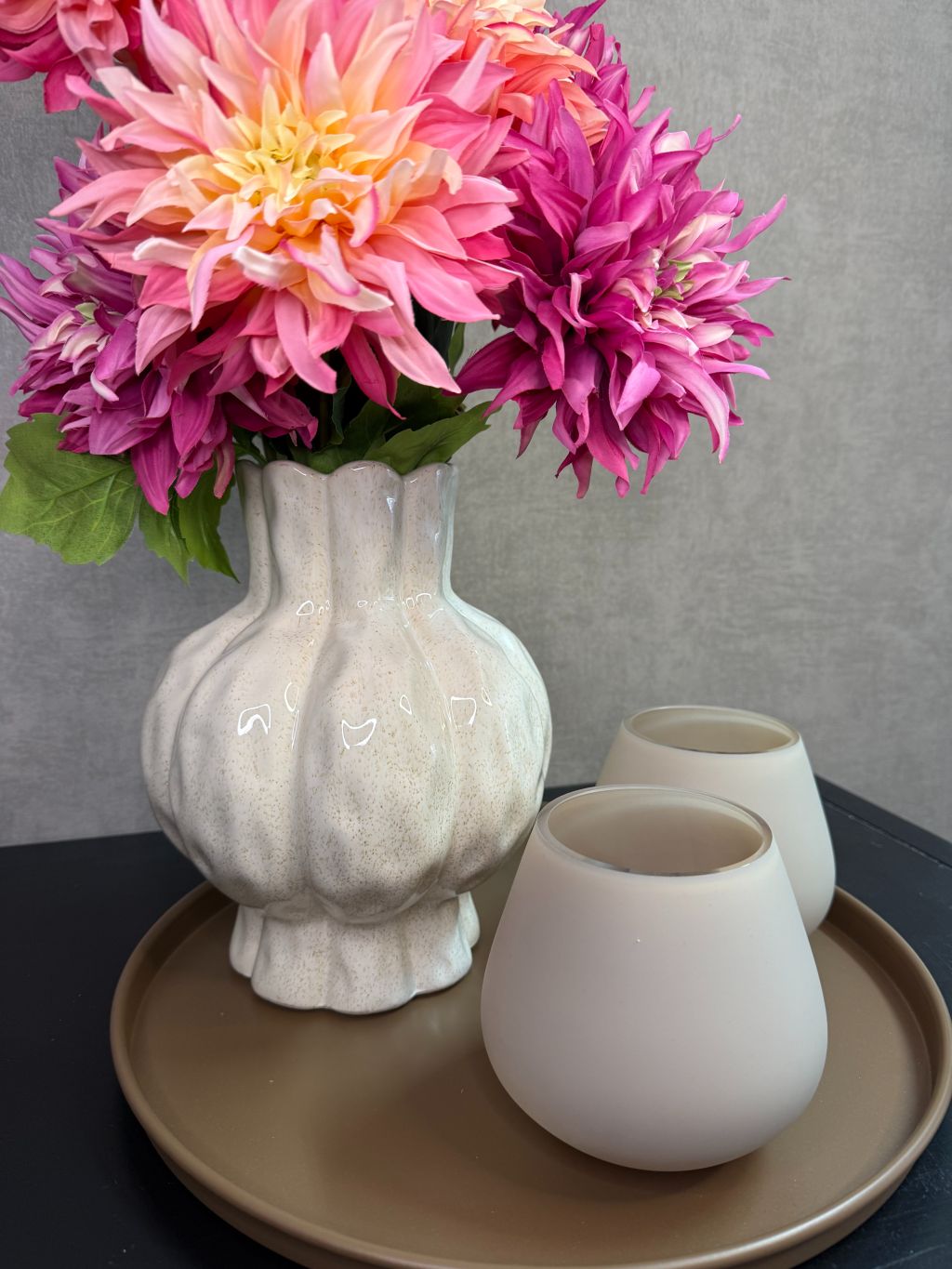 Tafel decoratie set met beige vaas, roze en perzikkleurige bloemen en twee waxinelichthouders op een rond bruin dienblad.