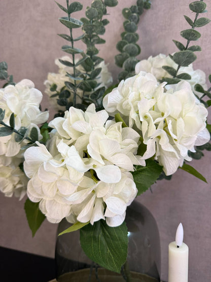 Goudkleurig dienblad met vaas met witte hortensia’s en eucalyptus, zwarte kandelaars, glazen waxinelichthouders en amberblokjes.