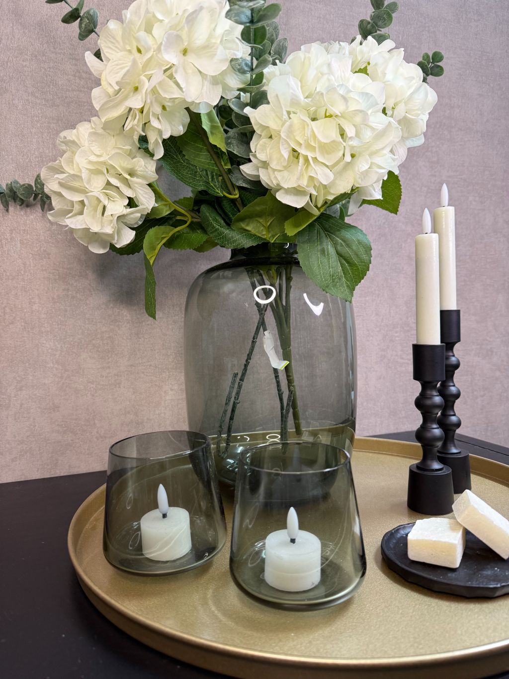 Goudkleurig dienblad met vaas met witte hortensia’s en eucalyptus, zwarte kandelaars, glazen waxinelichthouders en amberblokjes.