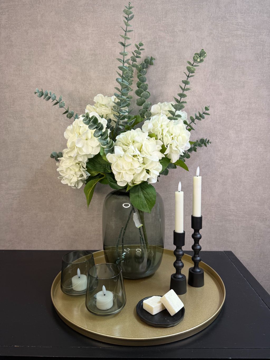 Goudkleurig dienblad met vaas met witte hortensia’s en eucalyptus, zwarte kandelaars, glazen waxinelichthouders en amberblokjes.