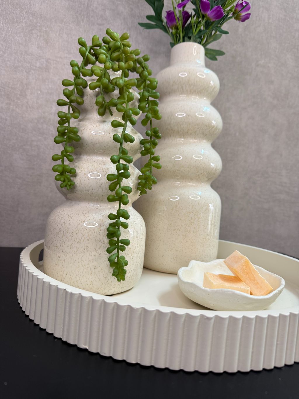 Tafel decoratie set met crèmekleurige vazen, paarse bloemen, groene hangplant en amberblokjes op een rond dienblad.