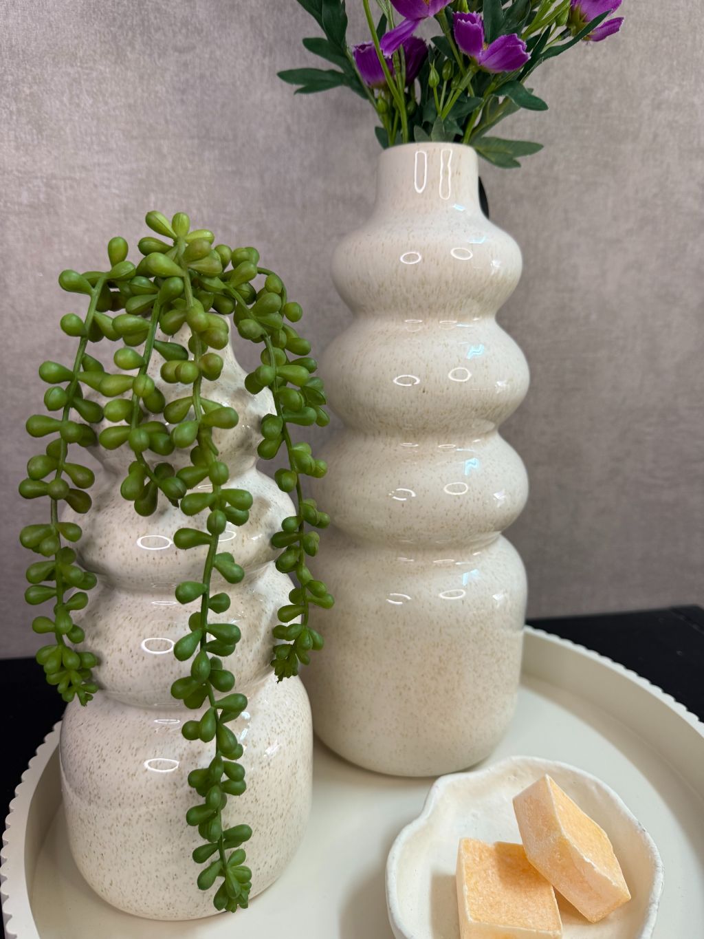 Tafel decoratie set met crèmekleurige vazen, paarse bloemen, groene hangplant en amberblokjes op een rond dienblad.