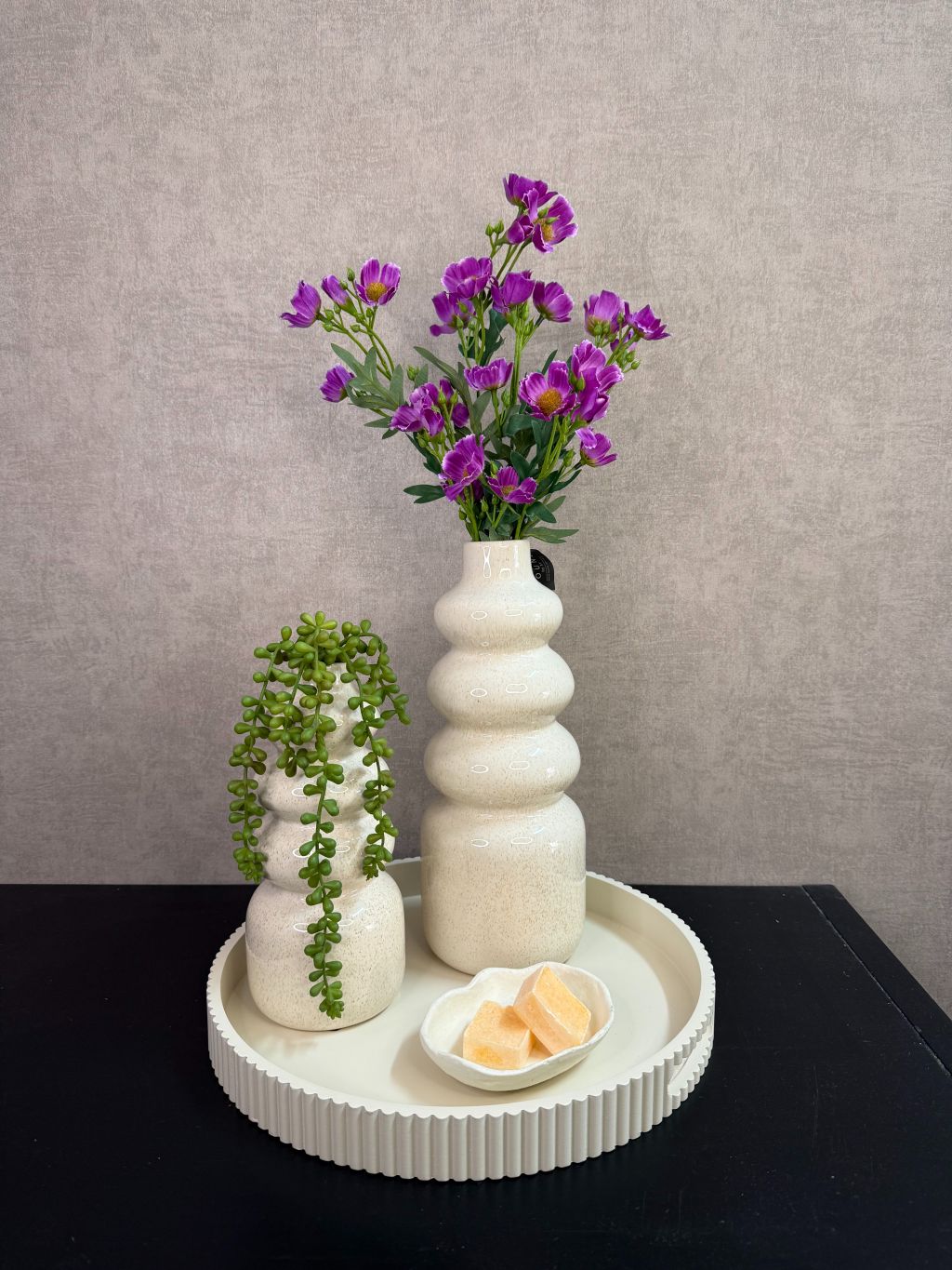 Tafel decoratie set met crèmekleurige vazen, paarse bloemen, groene hangplant en amberblokjes op een rond dienblad.