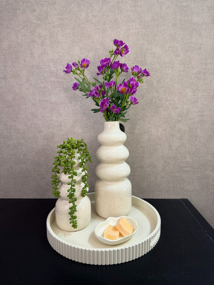 Tafel decoratie set met crèmekleurige vazen, paarse bloemen, groene hangplant en amberblokjes op een rond dienblad.