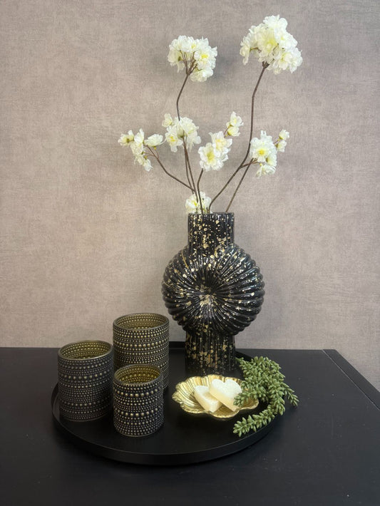 Tafel decoratie set Nelle – zwarte vaas met witte bloemen, goudkleurige schaal met amberblokjes en zwarte waxinelichthouders op rond dienblad.