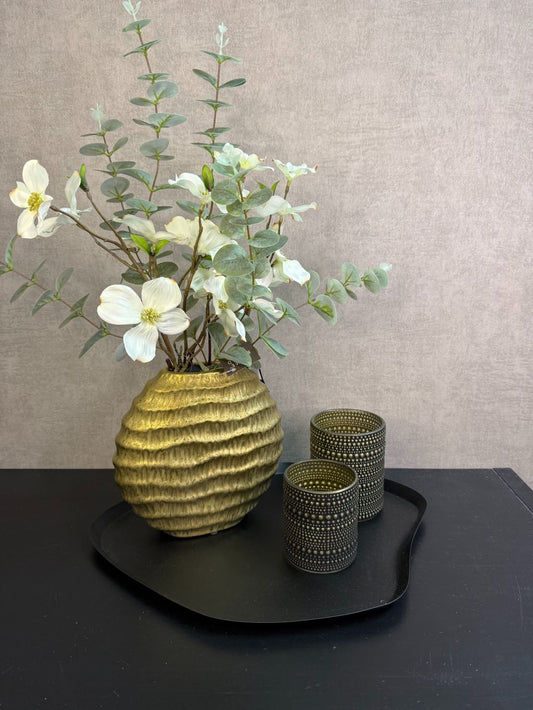 Zwart metalen dienblad met goudkleurige vaas met witte kunstbloemen en twee goud-zwarte waxinelichthouders op een zwarte tafel.