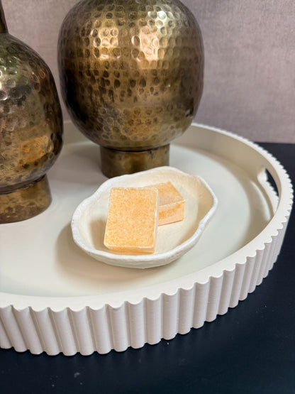 Ronde beige tray met twee goudkleurige vazen met groene kunstbloemen en een schaaltje met amberblokjes op een zwarte tafel.