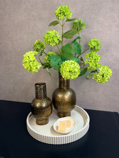 Ronde beige tray met twee goudkleurige vazen met groene kunstbloemen en een schaaltje met amberblokjes op een zwarte tafel.