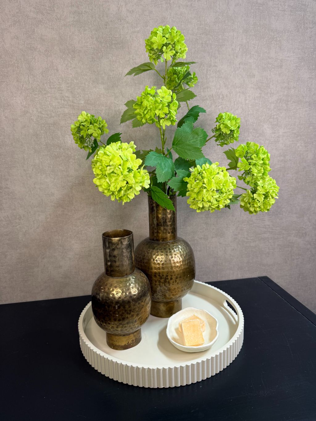 Ronde beige tray met twee goudkleurige vazen met groene kunstbloemen en een schaaltje met amberblokjes op een zwarte tafel.