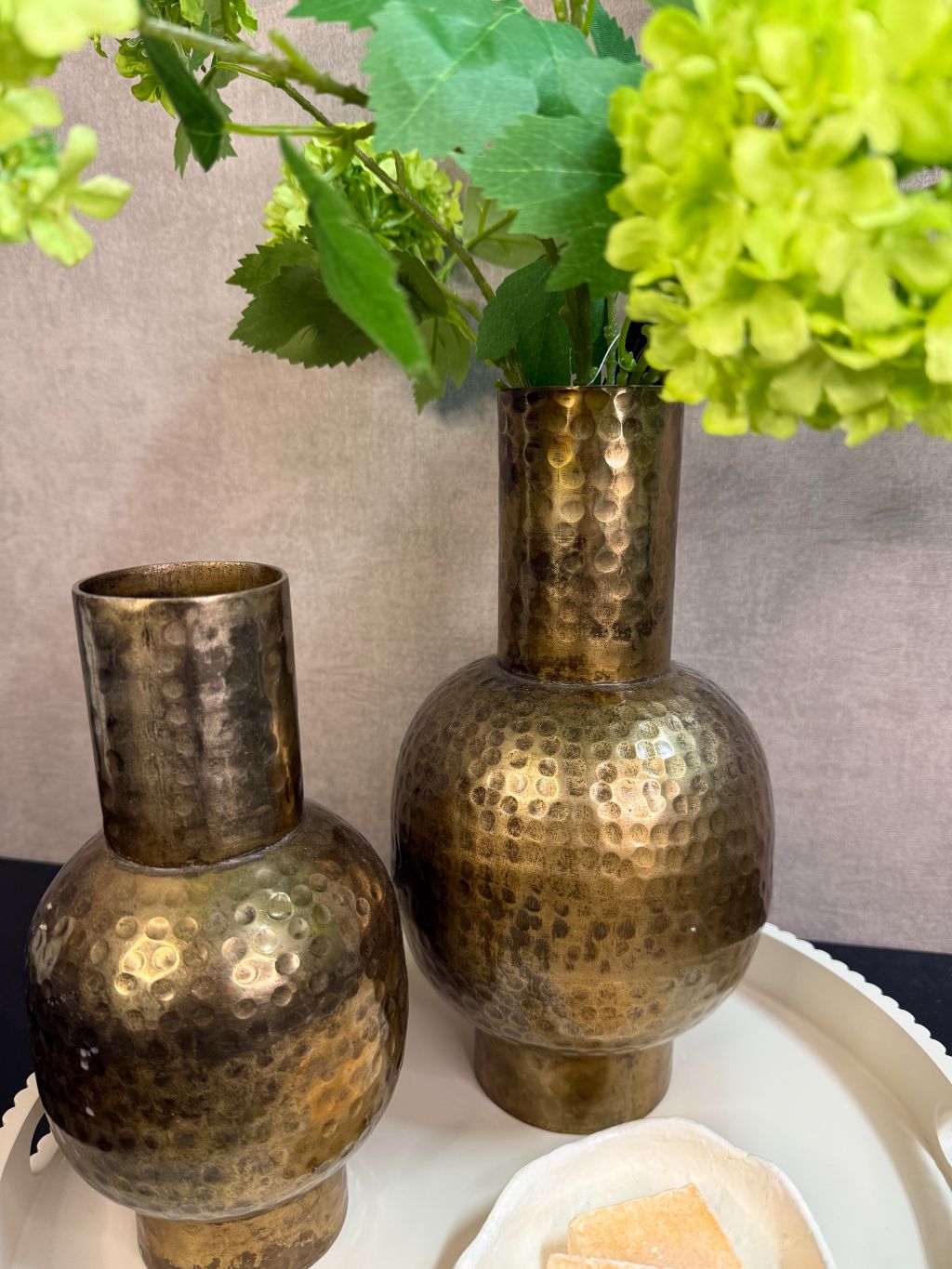 Ronde beige tray met twee goudkleurige vazen met groene kunstbloemen en een schaaltje met amberblokjes op een zwarte tafel.