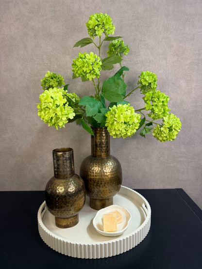 Ronde beige tray met twee goudkleurige vazen met groene kunstbloemen en een schaaltje met amberblokjes op een zwarte tafel.