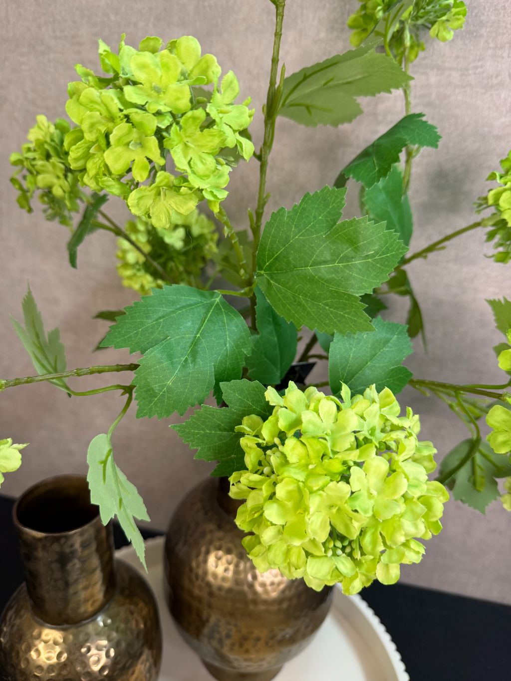 Ronde beige tray met twee goudkleurige vazen met groene kunstbloemen en een schaaltje met amberblokjes op een zwarte tafel.