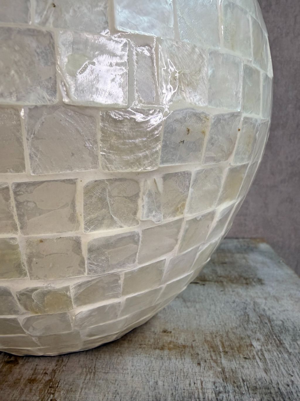 Ronde vaas met schelpmozaïek in crèmekleur, met smalle hals, geplaatst op een houten ondergrond tegen een neutrale achtergrond.