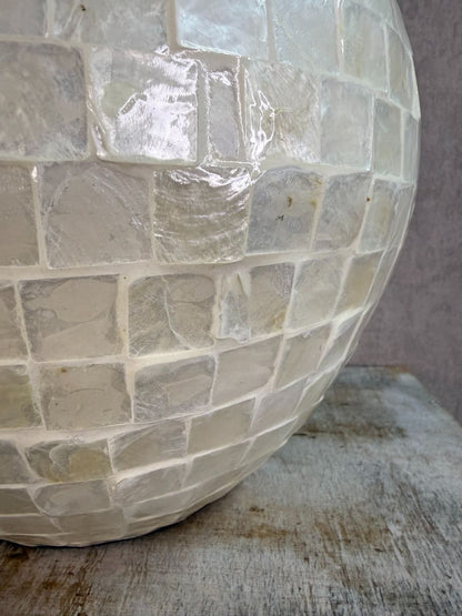 Ronde vaas met schelpmozaïek in crèmekleur, met smalle hals, geplaatst op een houten ondergrond tegen een neutrale achtergrond.