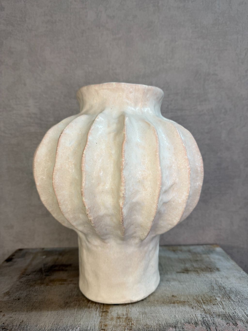 Beige keramieken vaas met organische ribstructuur op een houten ondergrond tegen een neutrale achtergrond.