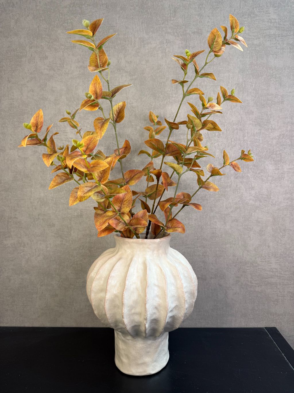 Beige keramieken vaas met organische ribstructuur op een houten ondergrond tegen een neutrale achtergrond met kunstbloemen.