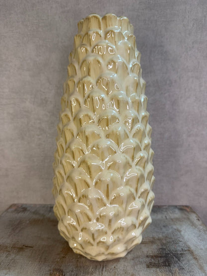 Vaas Pinecone beige – keramieken vaas met glanzende dennenappelstructuur op een houten ondergrond tegen een grijze muur.