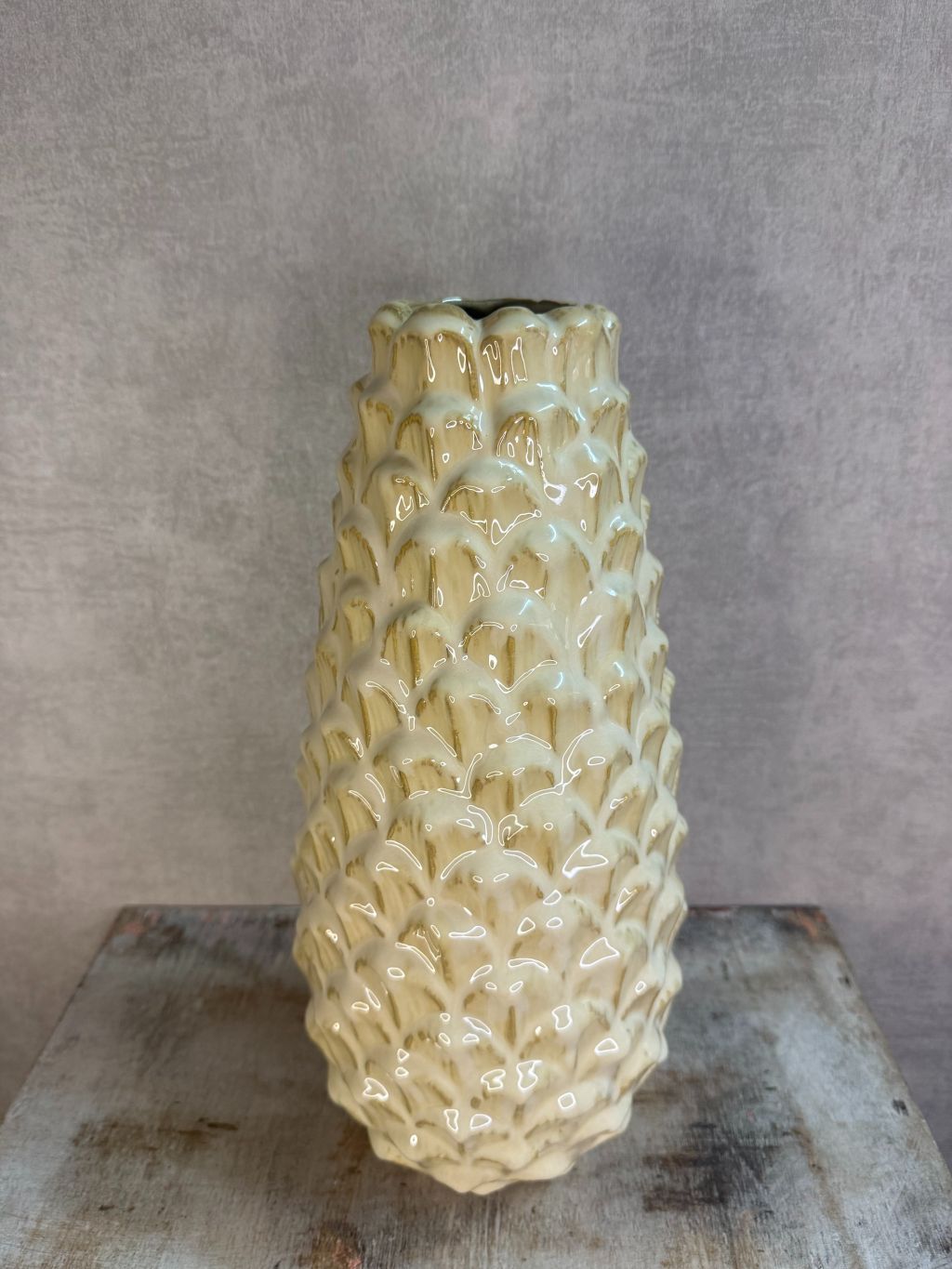 Vaas Pinecone beige – keramieken vaas met glanzende dennenappelstructuur op een houten ondergrond tegen een grijze muur.
