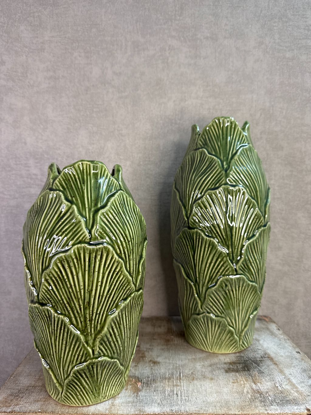 Groene keramieken vazen met bladstructuur in twee formaten op een houten ondergrond tegen een neutrale achtergrond.