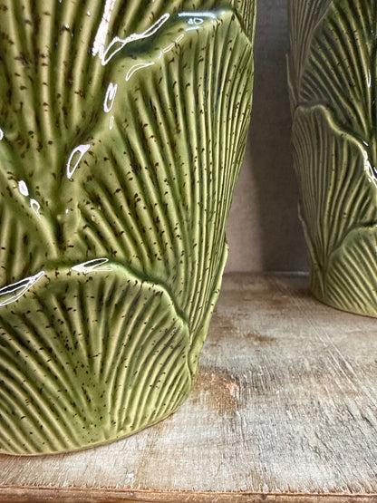 Groene keramieken vazen met bladstructuur in twee formaten op een houten ondergrond tegen een neutrale achtergrond.