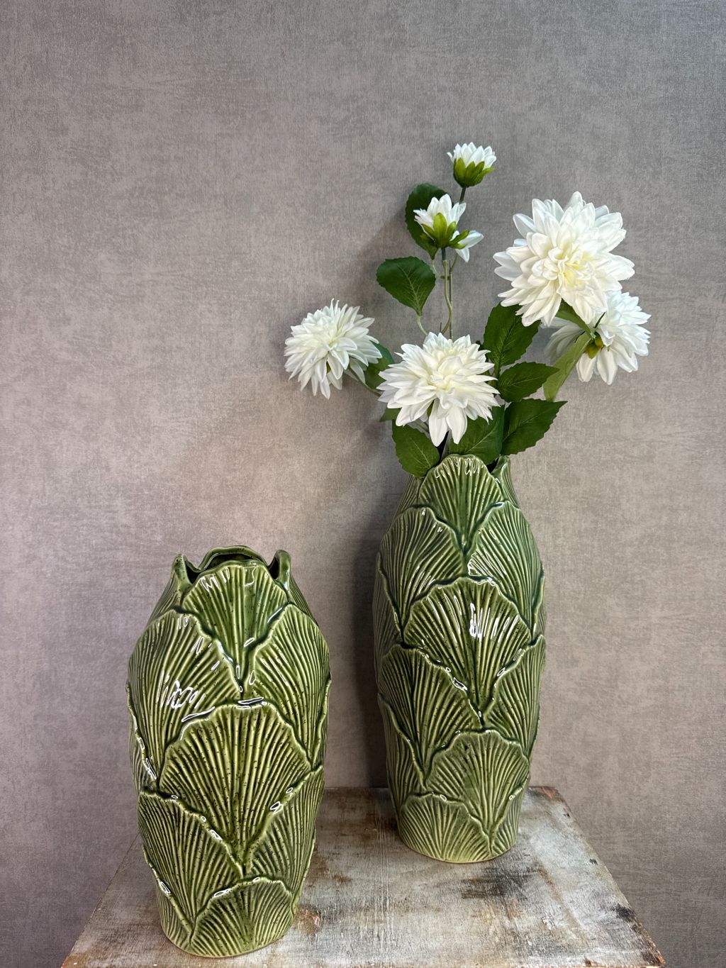 Groene keramieken vazen met bladstructuur in twee formaten op een houten ondergrond tegen een neutrale achtergrond.