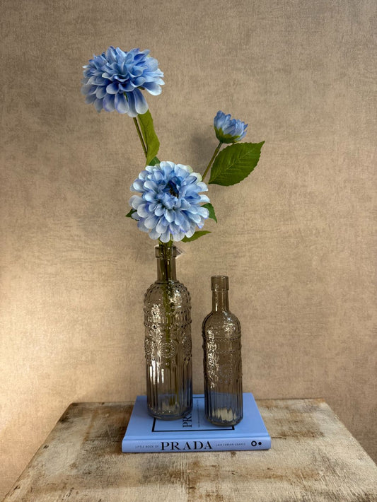Decoratieset met lichtblauw Prada-boek, twee rookglazen flesvazen en blauwe kunst dahlia tegen een warme beige achtergrond.
