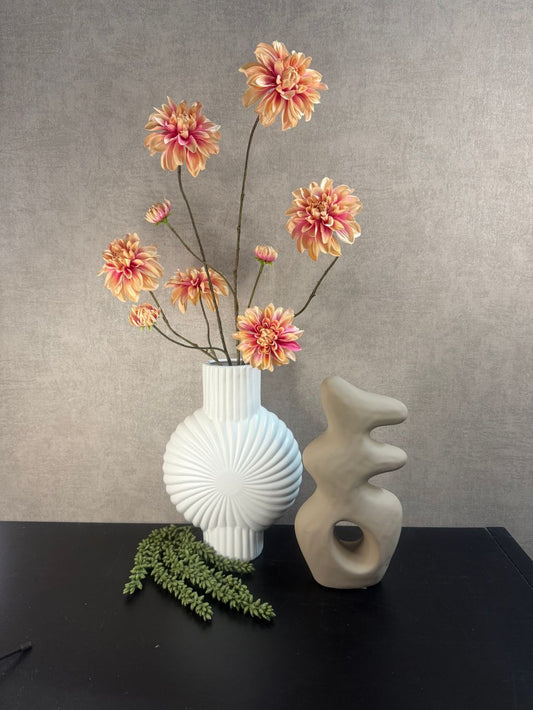 Witte vaas met roze dahlia’s, beige ornament en hangend kunstplantje op een zwarte ondergrond, gestyled als decoratie voor vensterbank of kast.