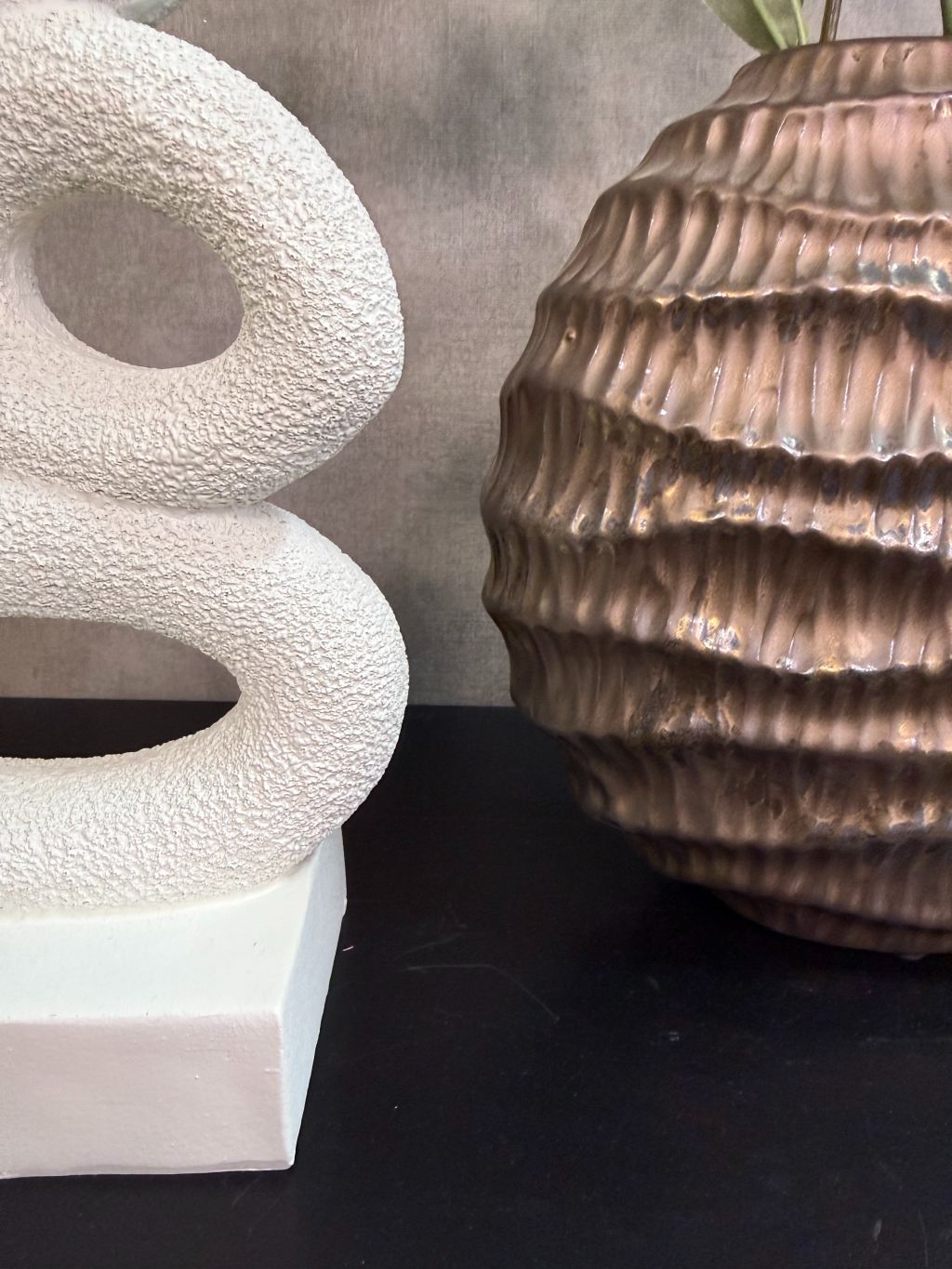 Bronzen keramieken vaas met witte bloesemtakken en groen blad naast een wit sculpturaal ornament op een zwarte tafel