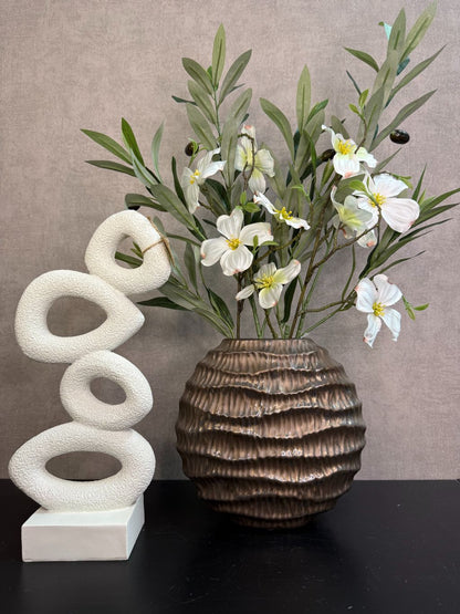 Bronzen keramieken vaas met witte bloesemtakken en groen blad naast een wit sculpturaal ornament op een zwarte tafel