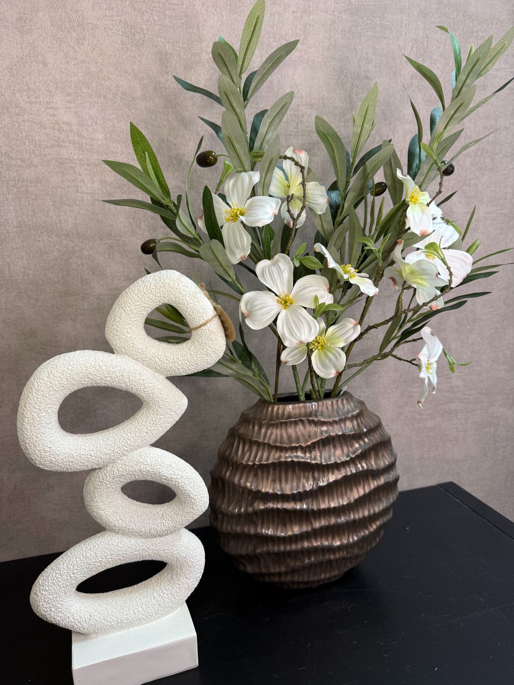 Bronzen keramieken vaas met witte bloesemtakken en groen blad naast een wit sculpturaal ornament op een zwarte tafel