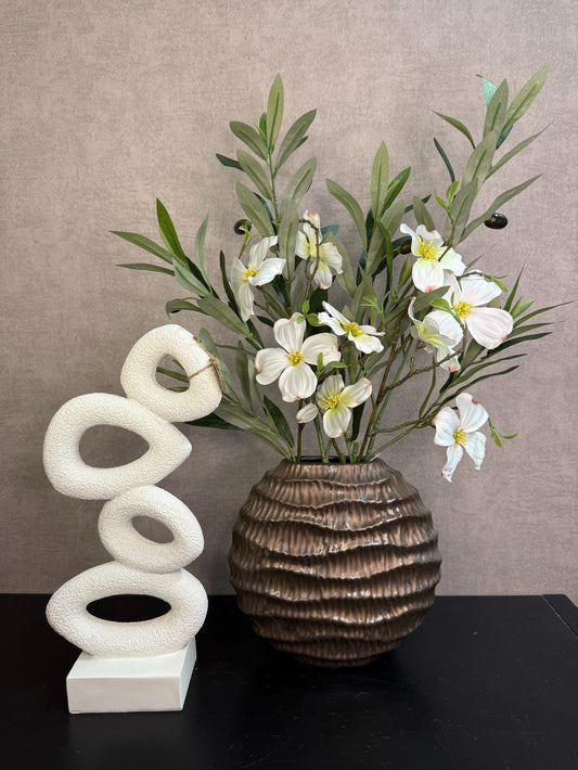 Bronzen keramieken vaas met witte bloesemtakken en groen blad naast een wit sculpturaal ornament op een zwarte tafel