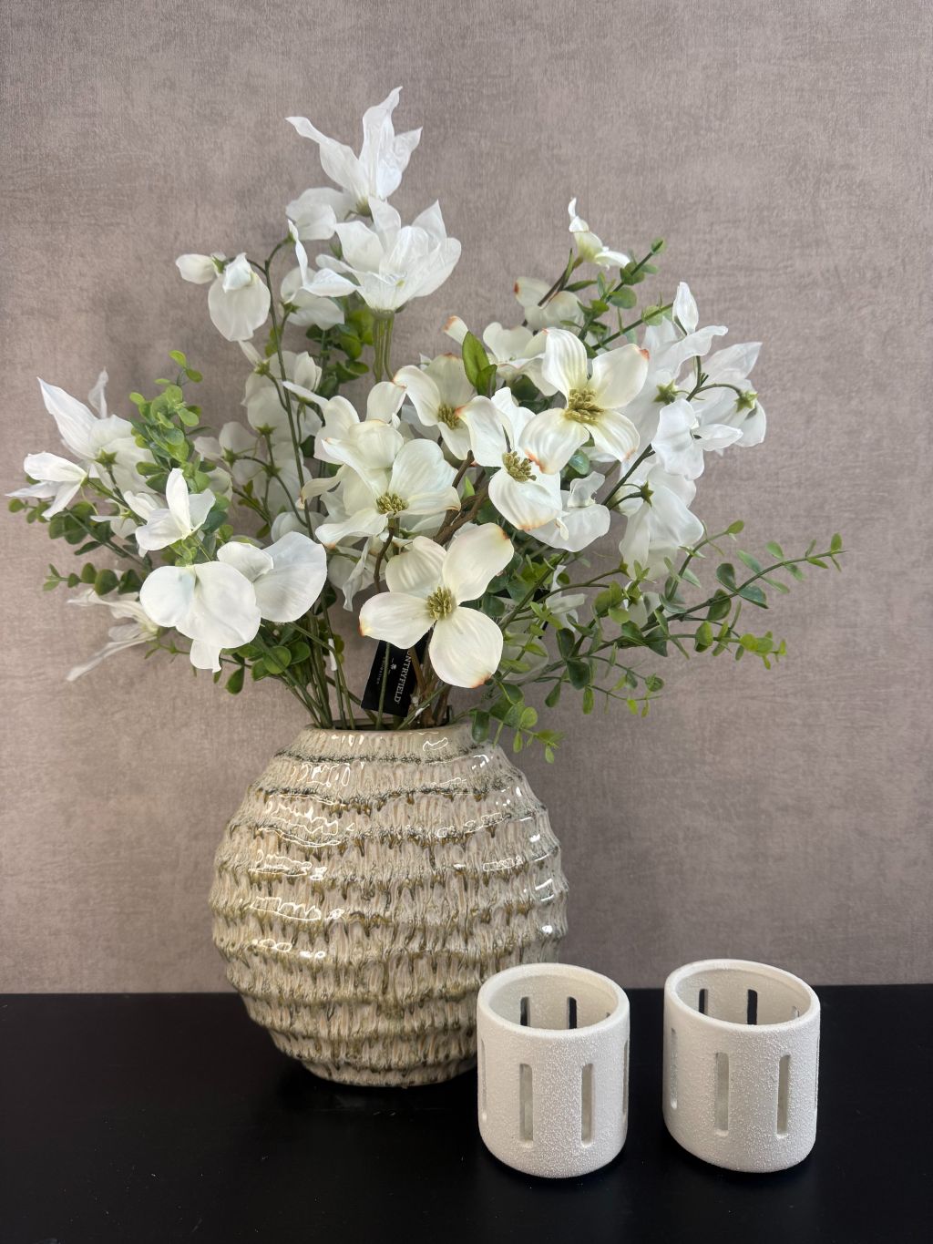 Vensterbank decoratieset met groene vaas gevuld met witte kunstbloemen en twee witte waxinelichthouders, op een zwarte ondergrond.