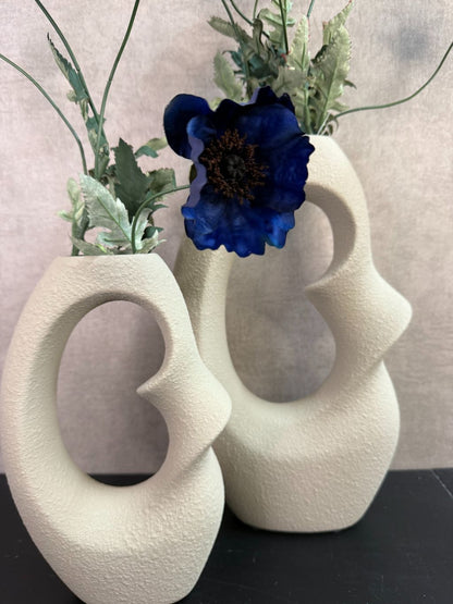 Beige design vaas met open vorm gecombineerd met donkerblauwe kunstbloemen op een houten ondergrond.