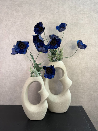 Beige design vaas met open vorm gecombineerd met donkerblauwe kunstbloemen op een houten ondergrond.