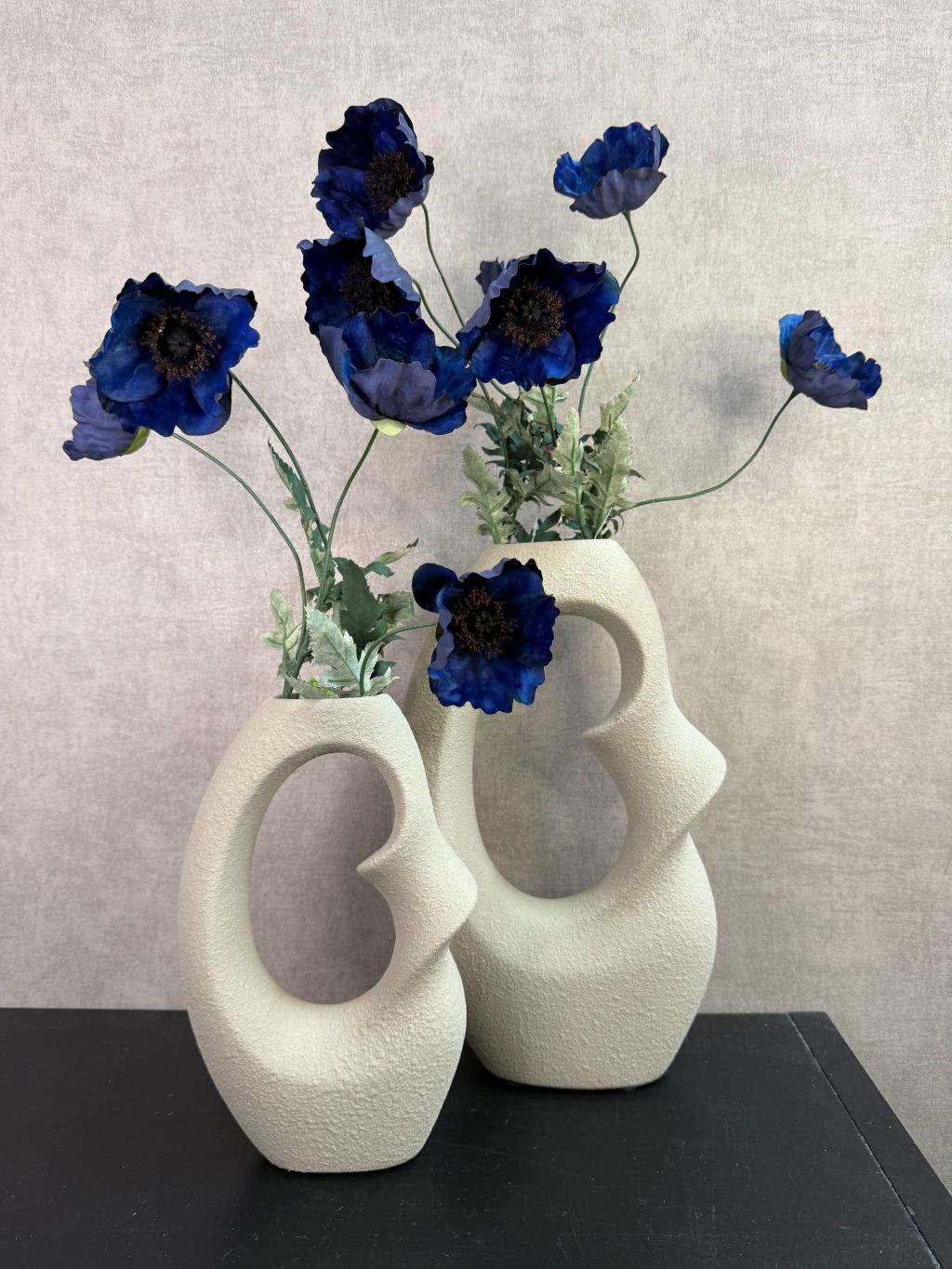 Beige design vaas met open vorm gecombineerd met donkerblauwe kunstbloemen op een houten ondergrond.