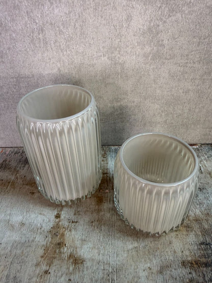 Twee beige waxinelichthouders van geribbeld glas in verschillende formaten op een houten ondergrond.