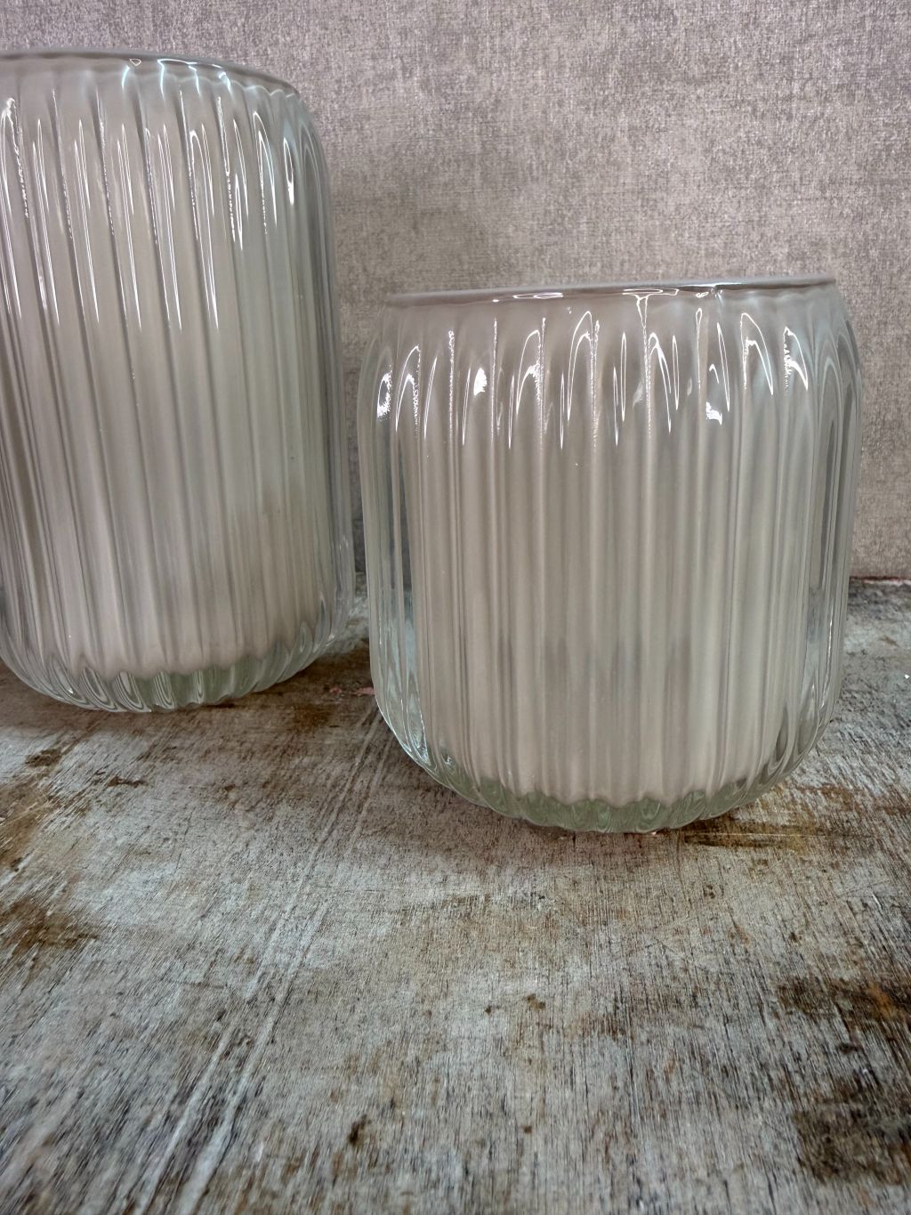 Twee beige waxinelichthouders van geribbeld glas in verschillende formaten op een houten ondergrond.