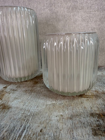 Twee beige waxinelichthouders van geribbeld glas in verschillende formaten op een houten ondergrond.