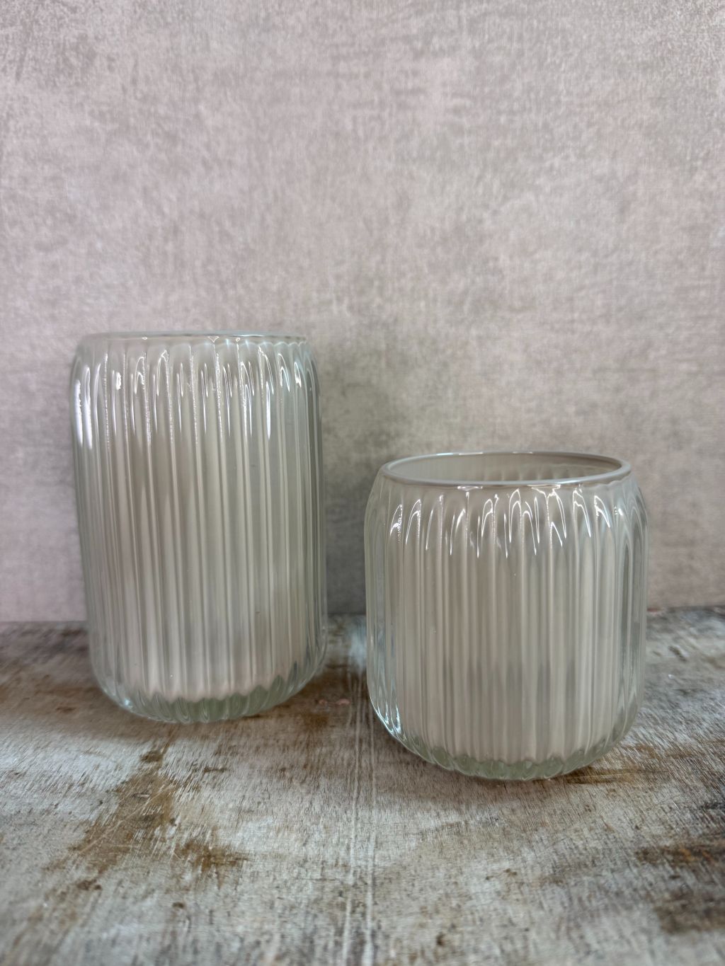 Twee beige waxinelichthouders van geribbeld glas in verschillende formaten op een houten ondergrond.