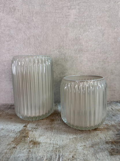 Twee beige waxinelichthouders van geribbeld glas in verschillende formaten op een houten ondergrond.